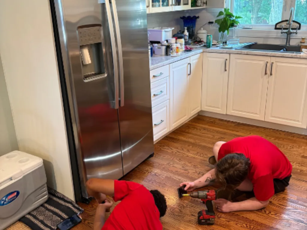 Two Appliance Shark technicians in red shirts working on the floor repairing a stainless steel side-by-side refrigerator freezer in a Prairie Village, Kansas home