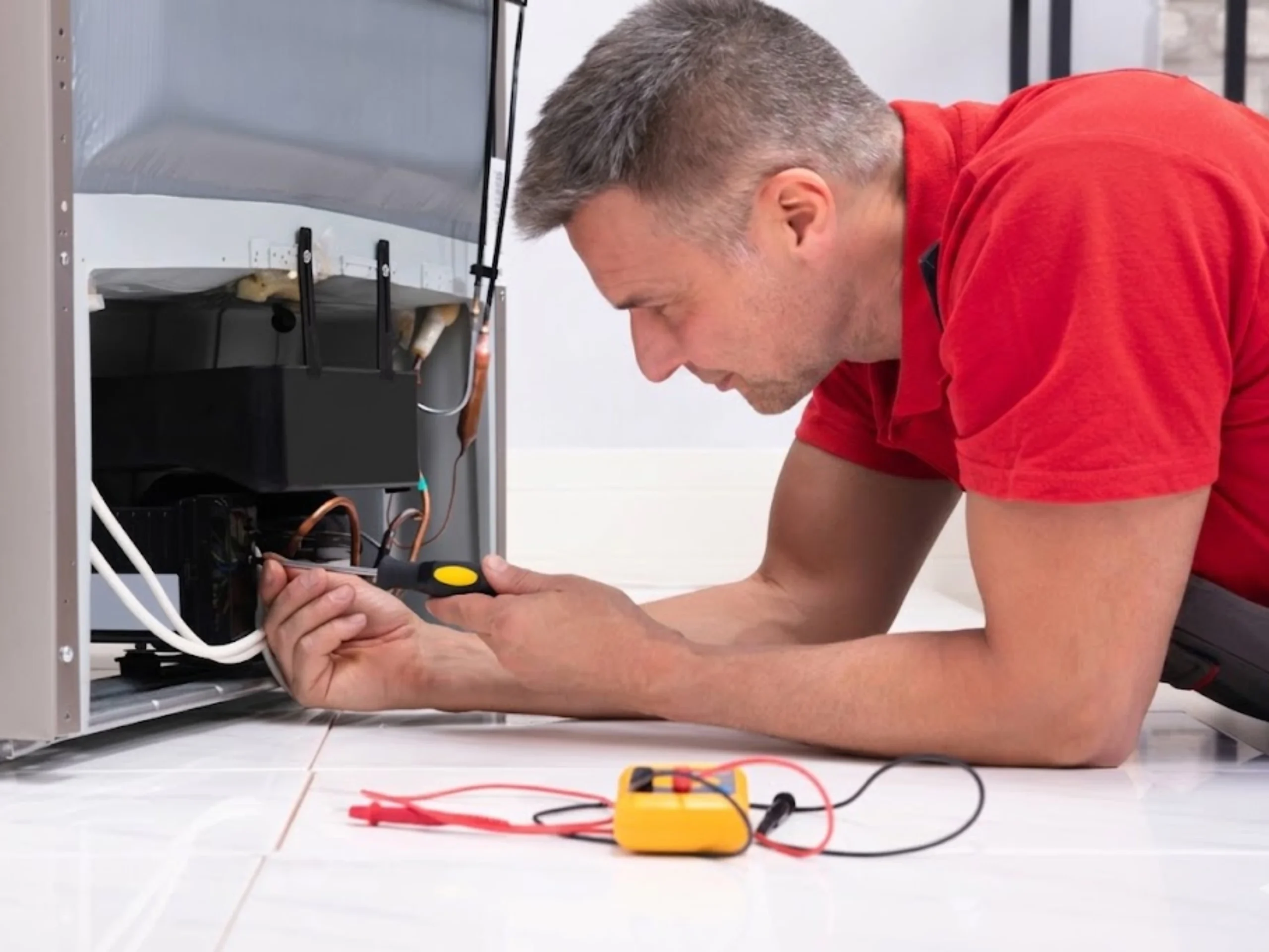 Appliance technician in a red shirt lying on the floor inspecting the internal components at the back of an ice maker, with a yellow multimeter on the floor beside him