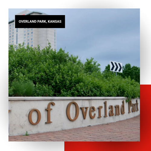 City of Overland Park, Kansas stone welcome sign surrounded by green trees with a hotel and cloudy sky in the background