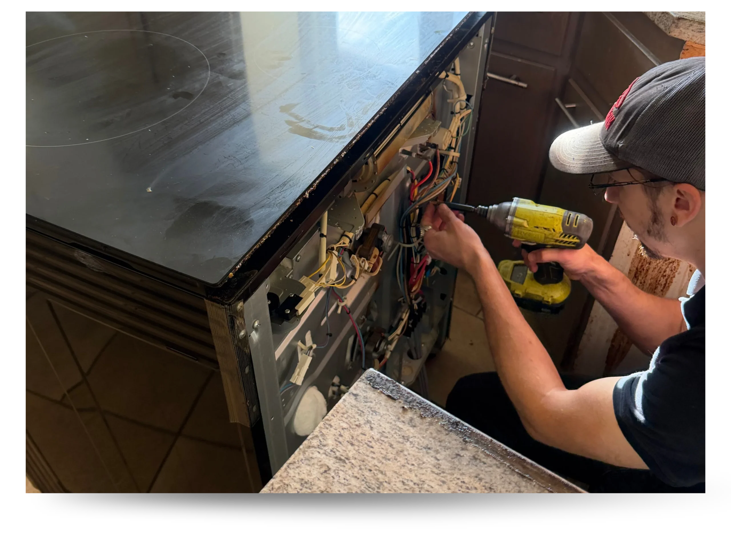 Appliance technician using a power drill to repair the internal wiring of a disassembled electric stove with glass cooktop in a kitchen