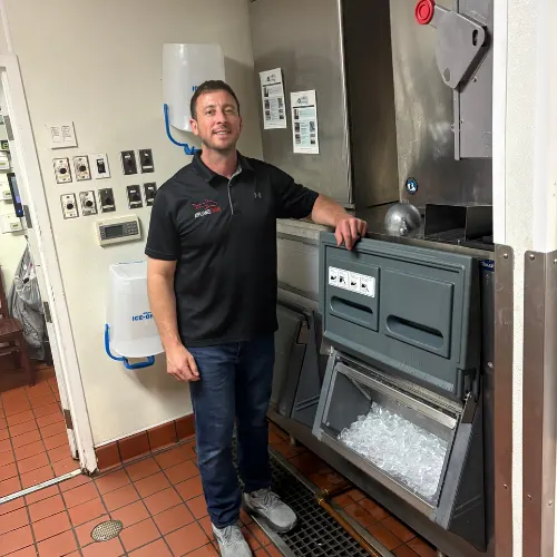 Appliance Shark technician in a black polo shirt smiling while standing next to a commercial ice machine in a commercial kitchen