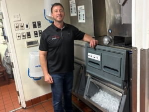 Smiling Appliance Shark technician in a black polo shirt standing next to a commercial ice machine in a commercial kitchen