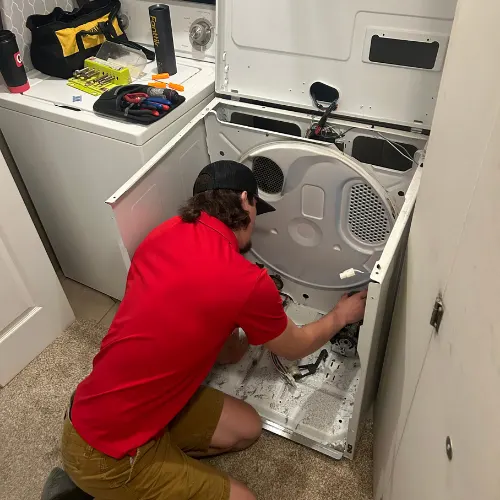 Appliance Shark technician in a red shirt kneeling to repair the interior of a disassembled dryer in a home laundry room