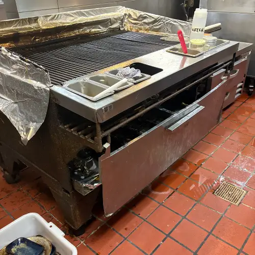 Large commercial charbroiler grill with open panels awaiting repair on a red tile floor in a restaurant kitchen