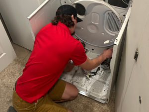 Appliance Shark technician in a red shirt kneeling to repair the interior of a disassembled residential dryer