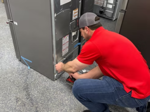 Technician in a red shirt and gray cap crouching down to repair the coil on a commercial refrigerator