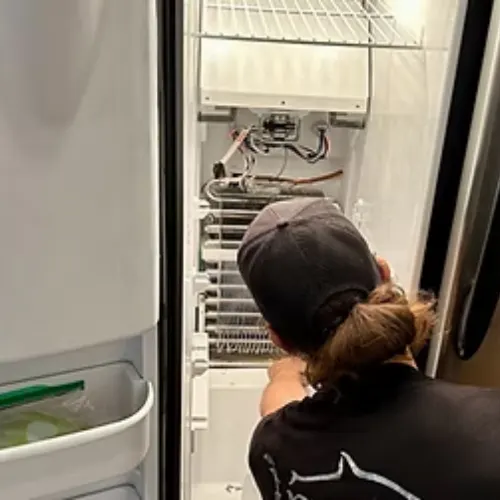 Appliance Shark technician in a black shirt and cap inspecting the evaporator coils and wiring inside an open upright freezer