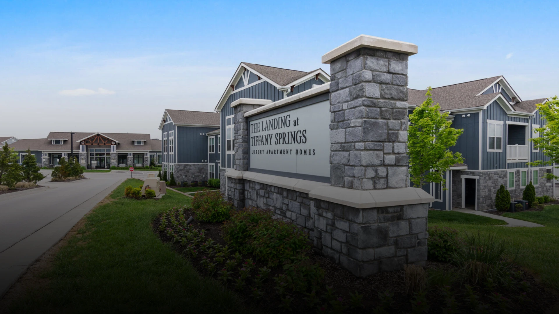 Stone entrance sign for The Landing at Tiffany Springs luxury apartment homes, with blue-gray apartment buildings in the background