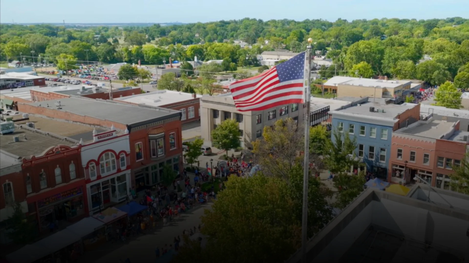 Aerial view of downtown Liberty, Missouri's historic square with a large American flag flying over brick buildings and a crowd gathered for a community event