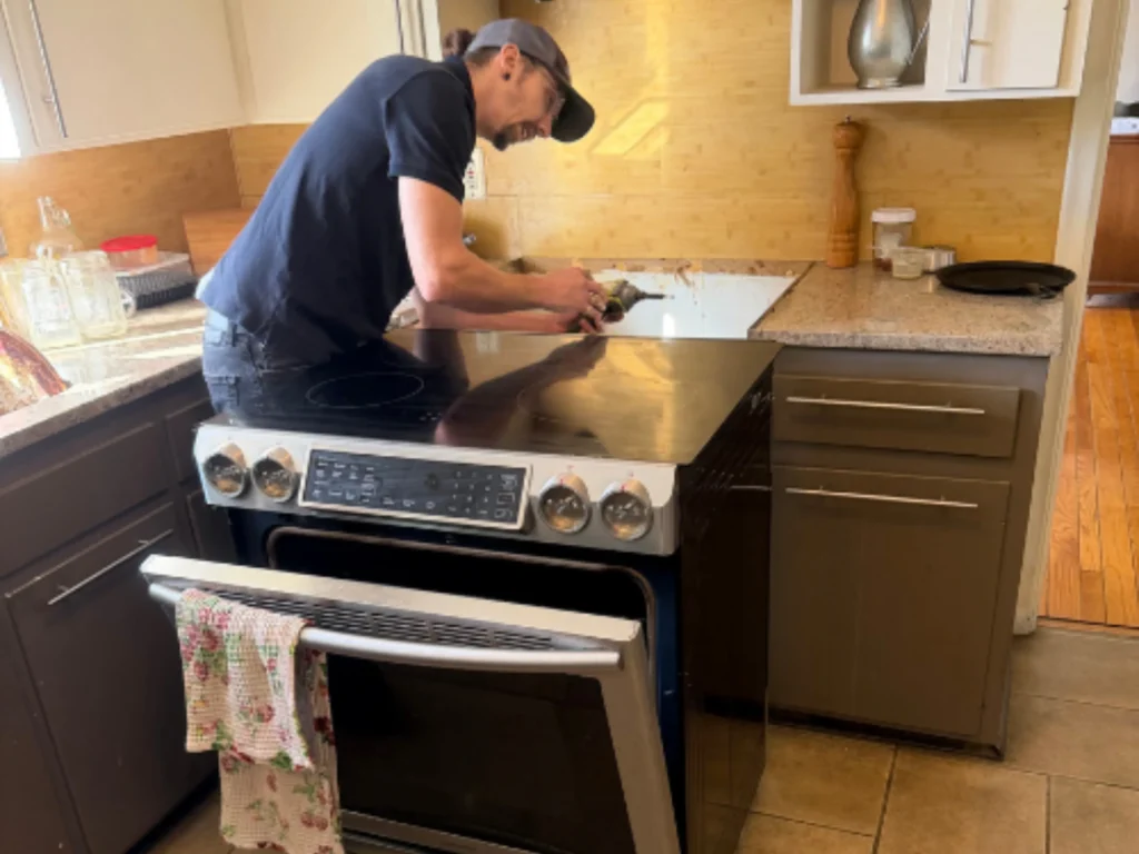 Technician adjusting the cooktop of a newly installed stainless steel slide-in electric range in a kitchen with tile flooring and brown cabinetry