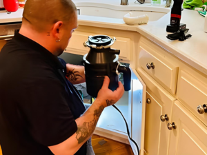 Tattooed technician in a black shirt installing or repairing a garbage disposal under a kitchen sink