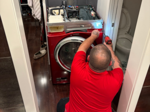 Appliance Shark technician in a red shirt kneeling to repair a red front-load washing machine in a home laundry area