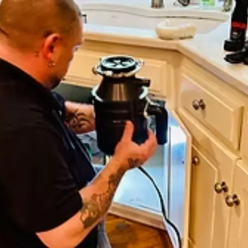 Tattooed technician in a black shirt replacing a garbage disposal unit under a kitchen sink cabinet