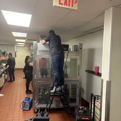 Technician standing on a ladder servicing a tall commercial glass-door refrigerator in a restaurant kitchen