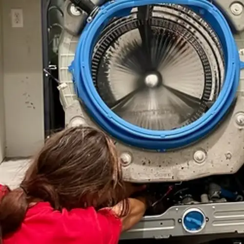 Appliance Shark technician in a red shirt leaning in to inspect the interior drum of a disassembled front-load washing machine