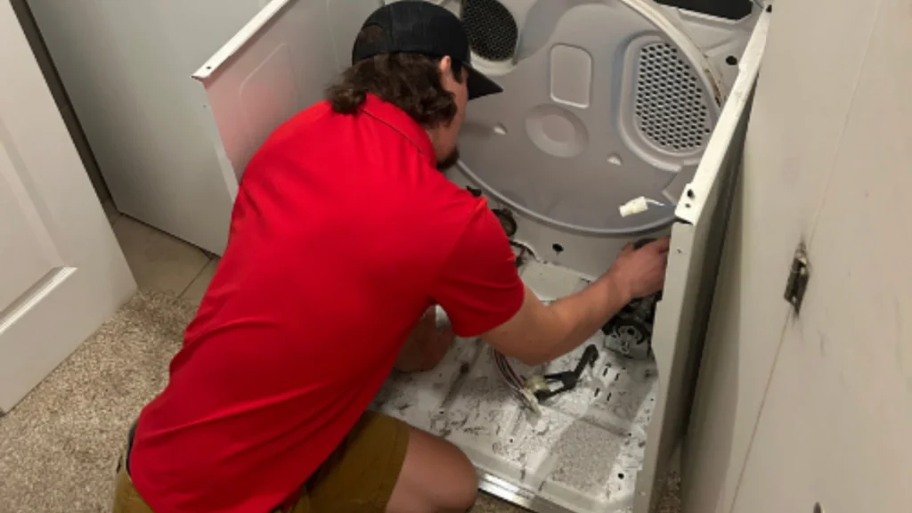 Appliance Shark technician repairing a dryer's internal components, kneeling beside the disassembled unit in a laundry closet